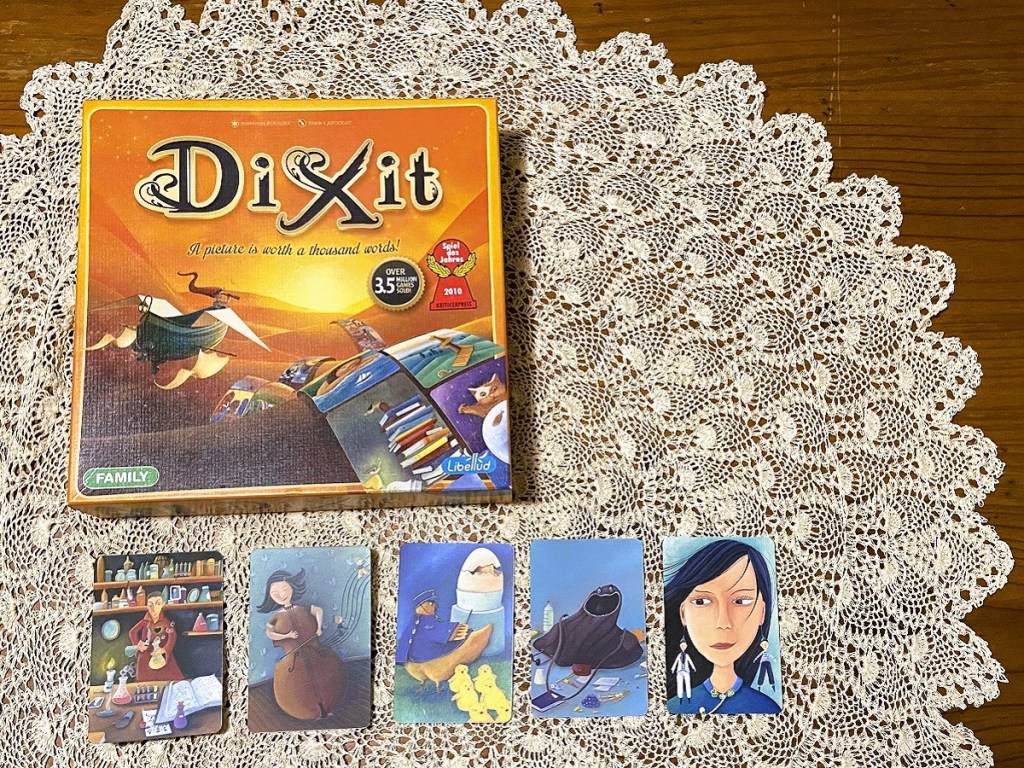 Image of DIXIT box with five cards face up, displayed on a Maltese lace tablecloth over a wood table