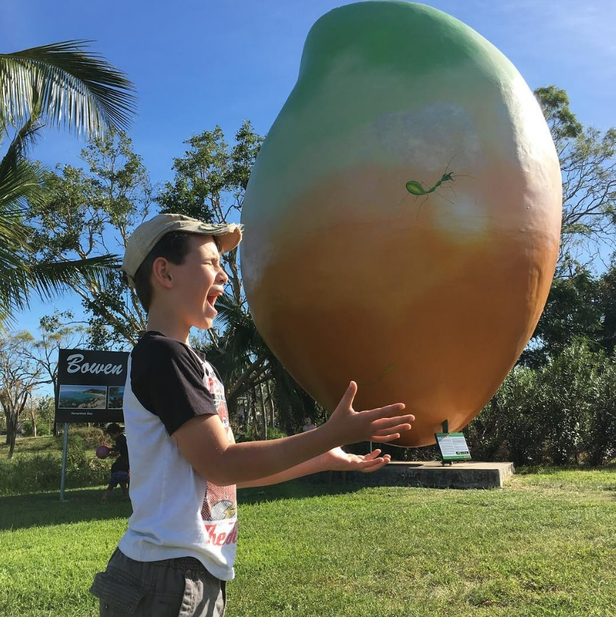 Image of young boy pretending to hold The Big Mango, a tourist site in Bowen Queensland. The image uses forced perspective to make the mango look closer. 
