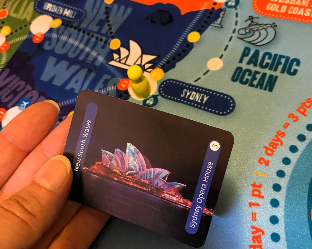 image of a hand holding a souvenir card from the tabletop game TRAVEL BUG, showing the Sydney Opera House