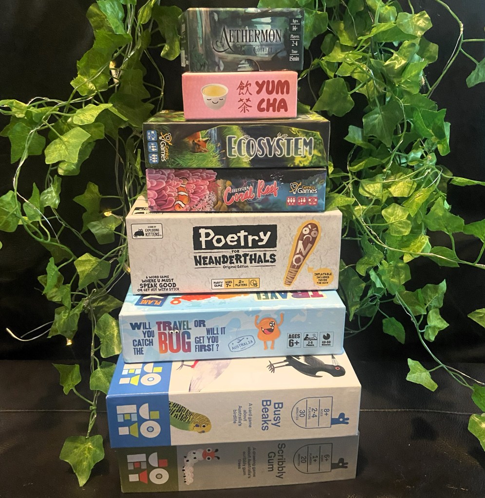 Image of a stack of 7 tabletop board games, including Aethermon Collect, Yum Cha, Ecosystem, Ecosystem Coral Reef, Poetry for Neanderthals, Travel Bug, Busy Beaks, and Scribbly Gum