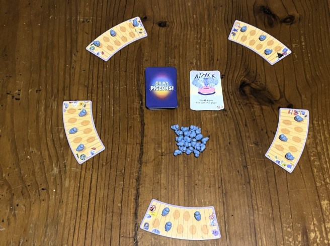 image of layout to start the tabletop game oh my pigeons showing five cardboard benches in a wide circle around the plastic pigeon meeple flock in the middle and a deck of face down cards with a face up discard pile to the right of the deck
