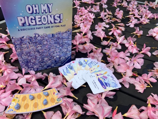 image of the box for the game oh my pigeons sitting on a table covered in pink flowers with cards spread out in front and a cardboard bench with pigeon meeples sitting on it