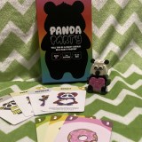 Photo image of the tabletop card game Panda Party set against a material backdrop of green and white zig zag stripes, with cards from the game spread out in front of the game box. There is a LEGO panda with a pink love heart sitting next to the box.