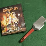 image of the box for tabletop game Burger Up on a green gaming cloth with a metal barbecue spatula next to it