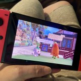 photo of nintendo switch held in hand while a person sits on the couch. The screen shows the video game Letters to Arralla with the main character, a turnip, asking what do they do.